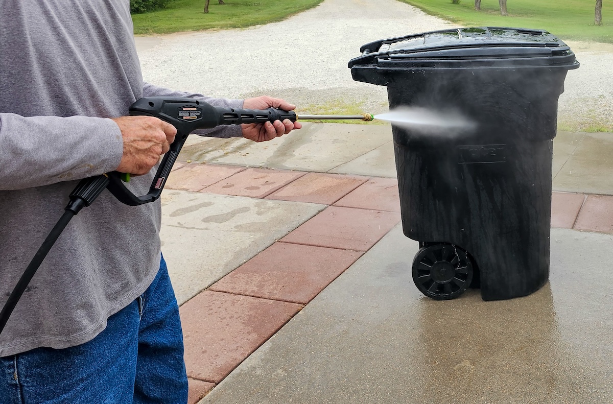 Man in grey shirt points pressure washer spray at a black outdoor trash bin.