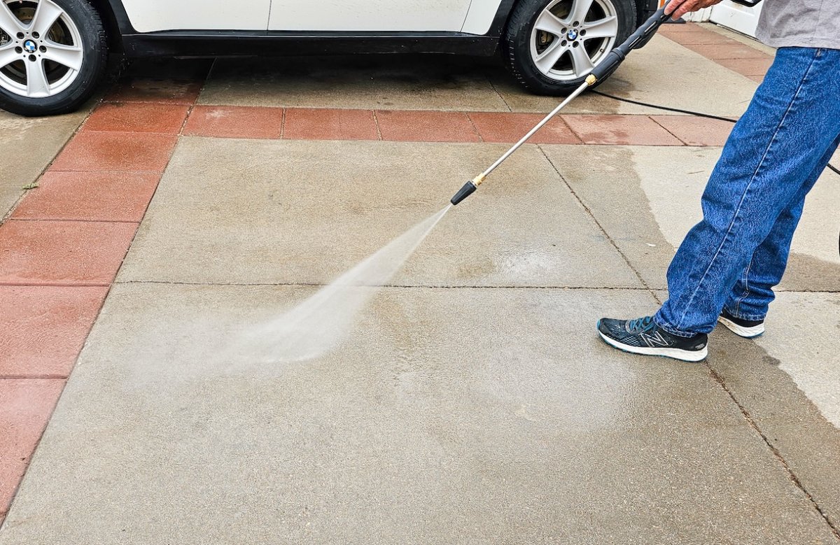 Shot of man from the waist down pressure washing a driveway with brick inset accents. A car visible in the background.