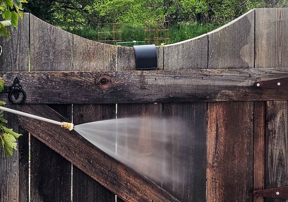 Pressure washing nozzle aims water at a weathered wooden fence.