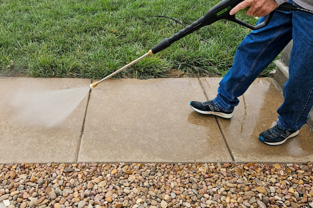 Man uses a pressure washer to clean sidewalk between grass and gravelly areas.