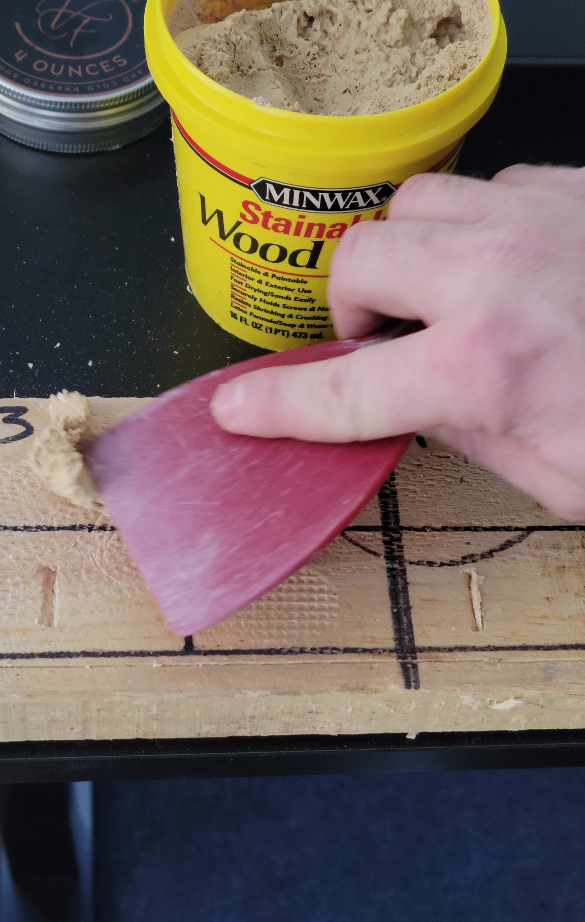 A person using a putty knife to scrape excess Minwax Stainable Wood Filler from a recently filled wood board.