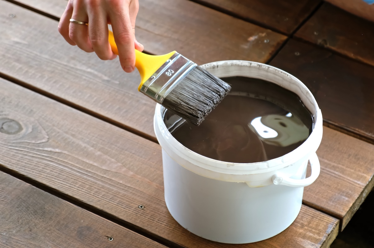 Person's hand dipping a brush into a bucket full of brown stain on a wooden deck.