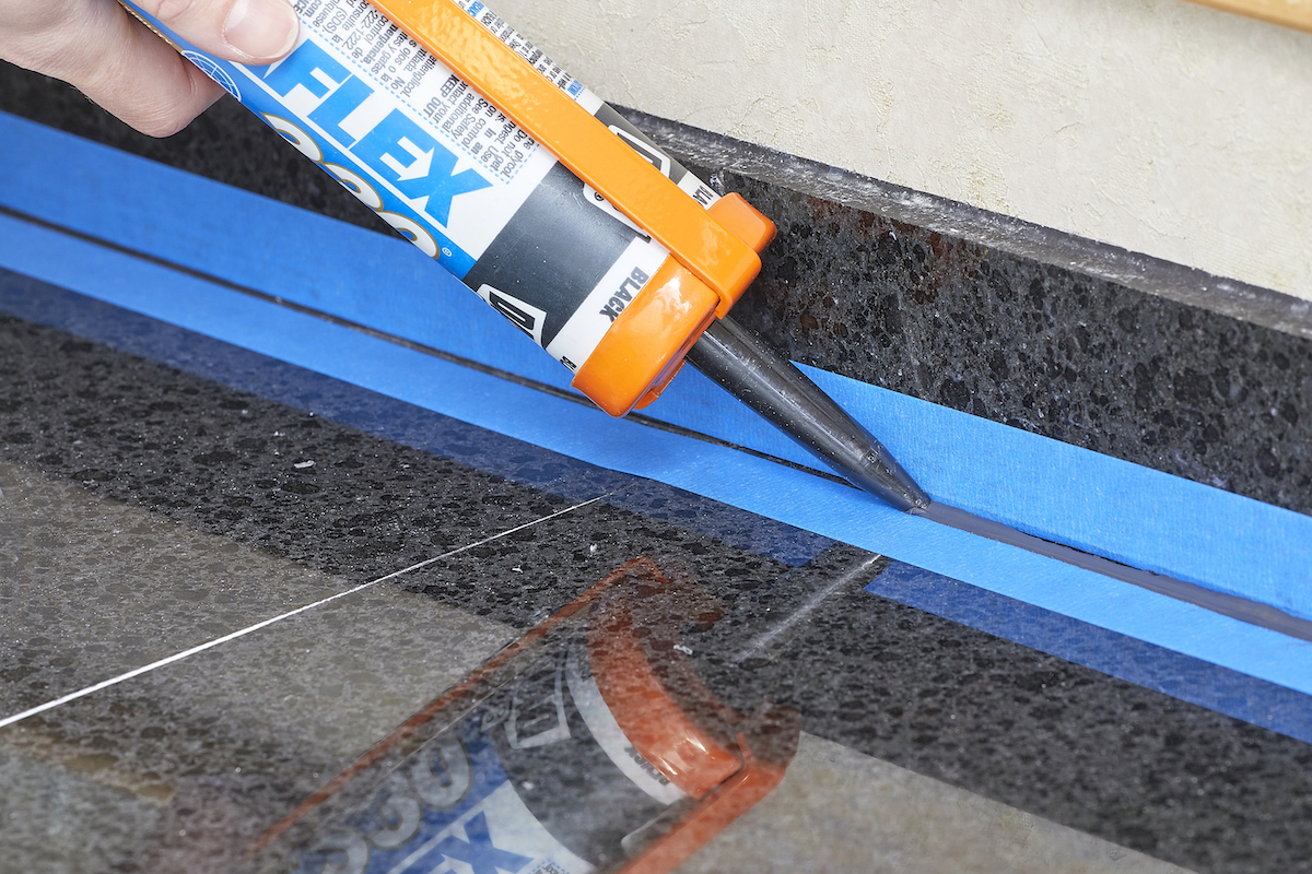 Person applies caulk to a gap between painter's tape rows on bathroom tile.