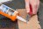 Woman uses an X-acto knife to cut the tip of the caulk tube; a piece of cardboard is underneath.