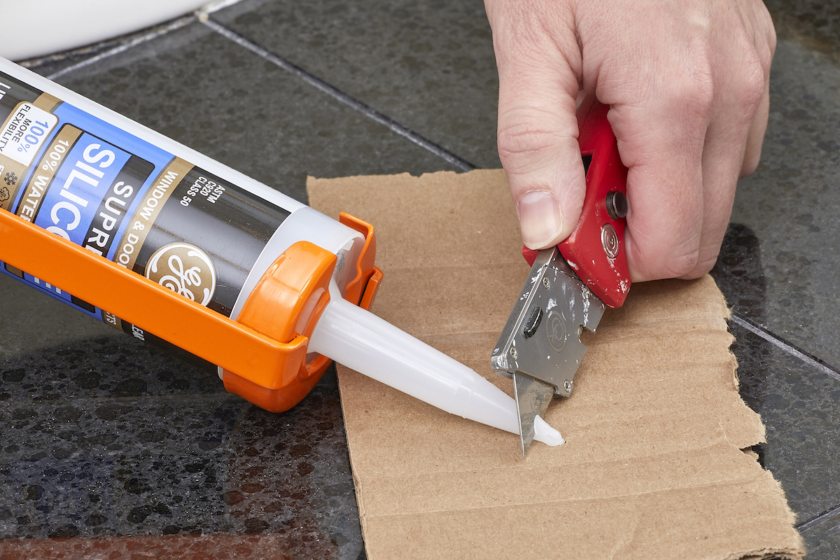 Woman uses an X-acto knife to cut the tip of the caulk tube; a piece of cardboard is underneath.