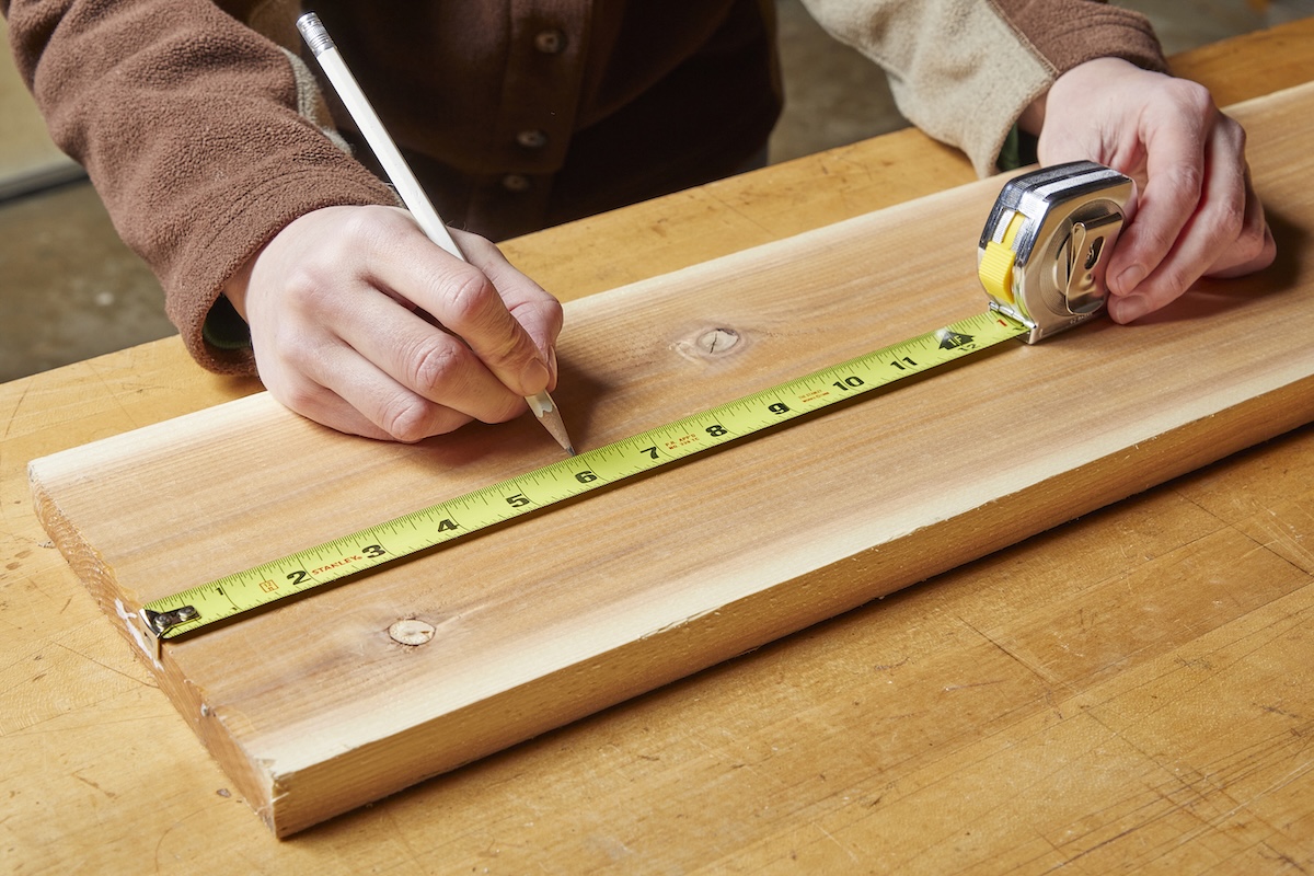 Person wearing brown jacket holding a pencil uses a tape measure to measure a board.