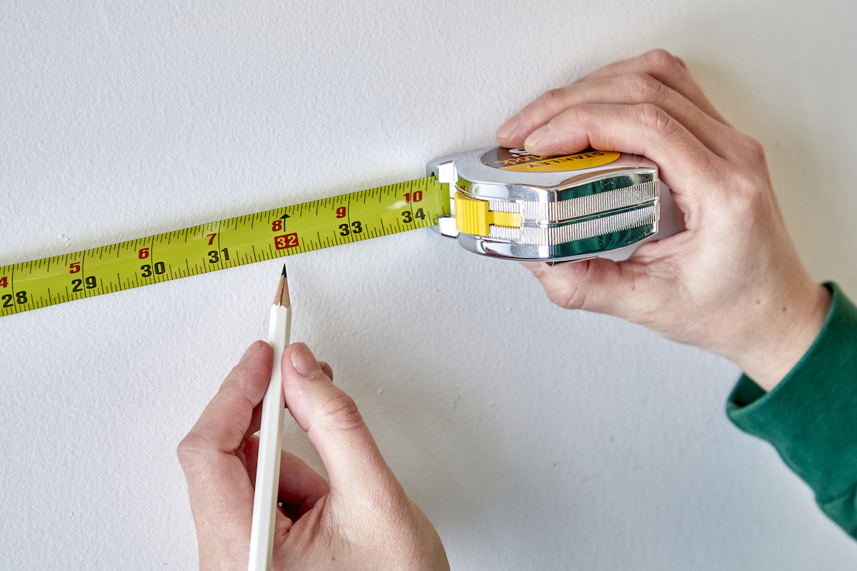 Woman points with a pencil to red rectangle on a tape measure that's stretched out on a wall.