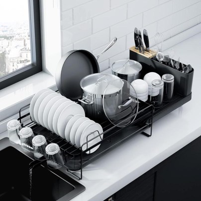 The Kitsure Dish-Drying Rack With Extendable Tray sitting on a kitchen counter next to a kitchen sink and loaded with drying dishes.
