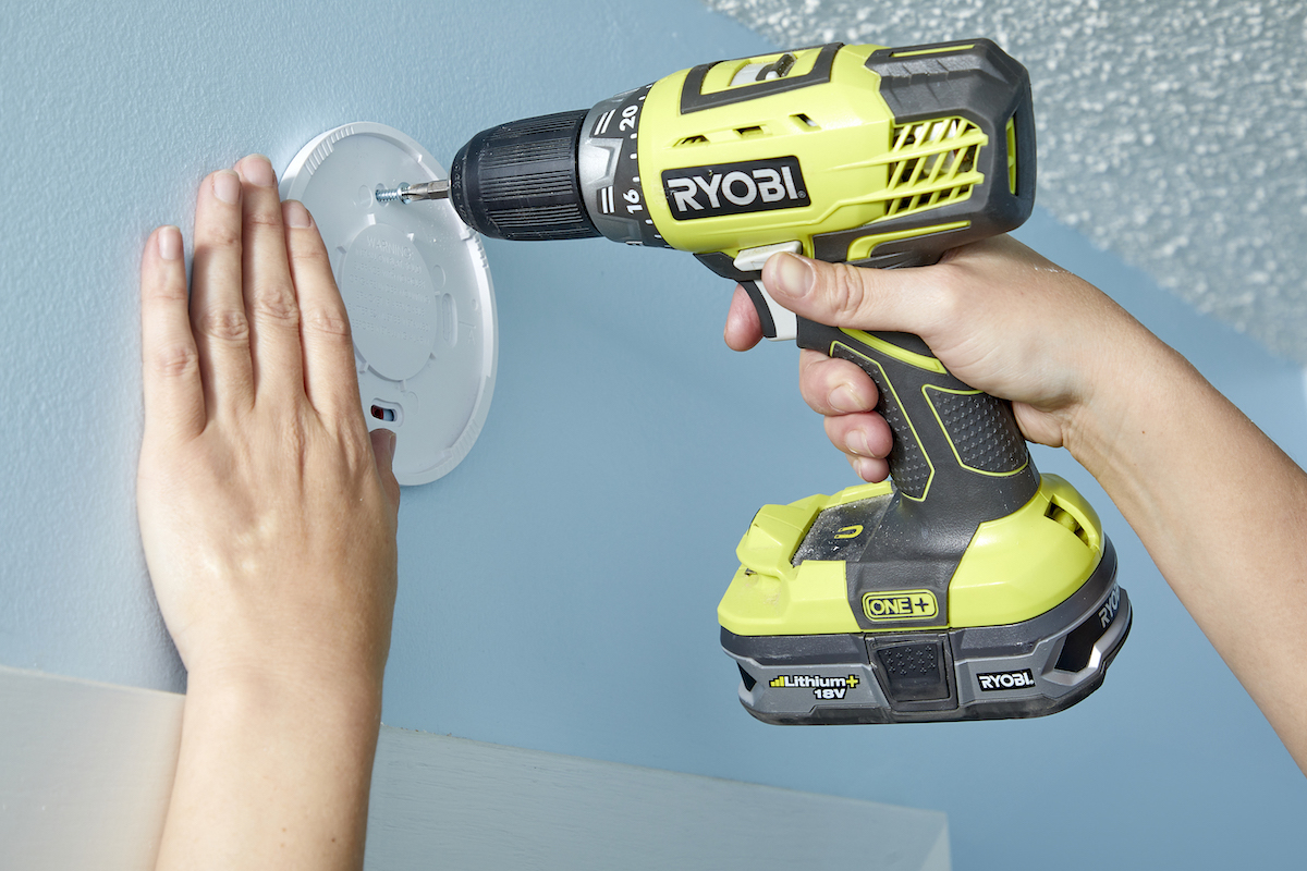 Woman drills screws through the mounting plate of a smoke detector into a wall.