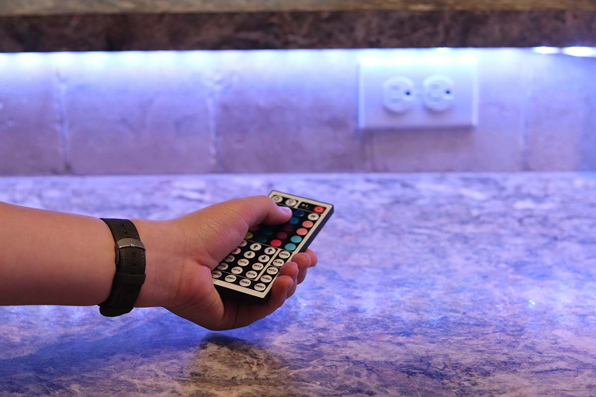 A person using a remote to turn on the best LED light strips that are installed under upper kitchen cupboards.