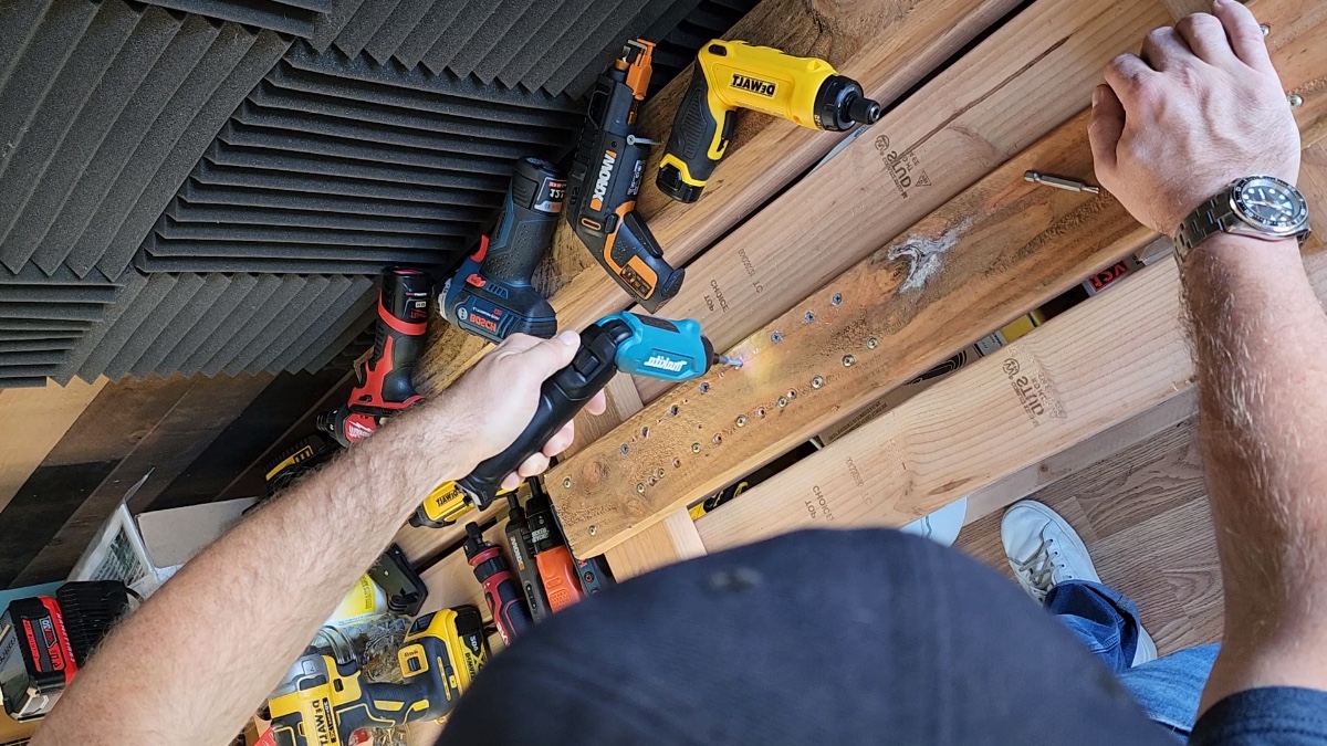 A person testing the best screwdriver option on a piece of wood with several other electric screwdrivers on a table in the background.