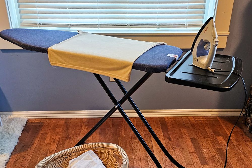 The Brabantia Adjustable Steam Rest Ironing Board set up in a laundry room with a towel on it, an iron, and a wicker basket with laundry on the floor.