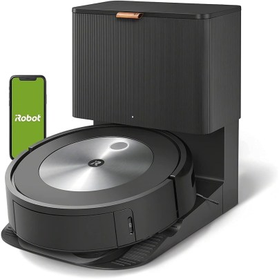 The iRobot Roomba j7+ Self-Emptying Robot Vacuum in its docking station with a phone next to it showing the iRobot app.