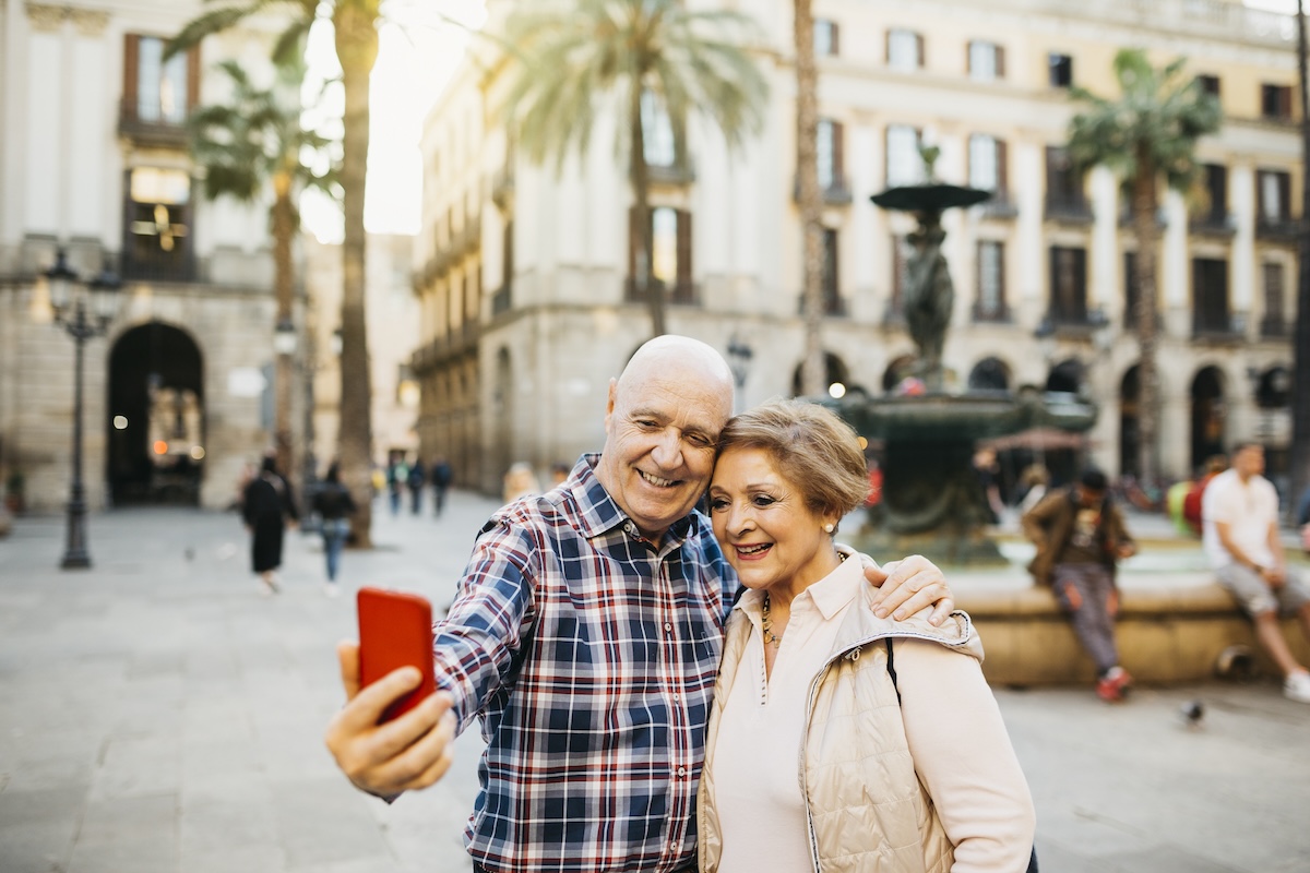 An older could poses for a selfie while in a historical city square on a vacation.