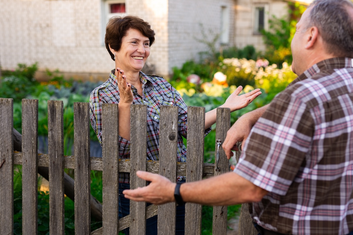 Two neighbors greet each other from across a fence.