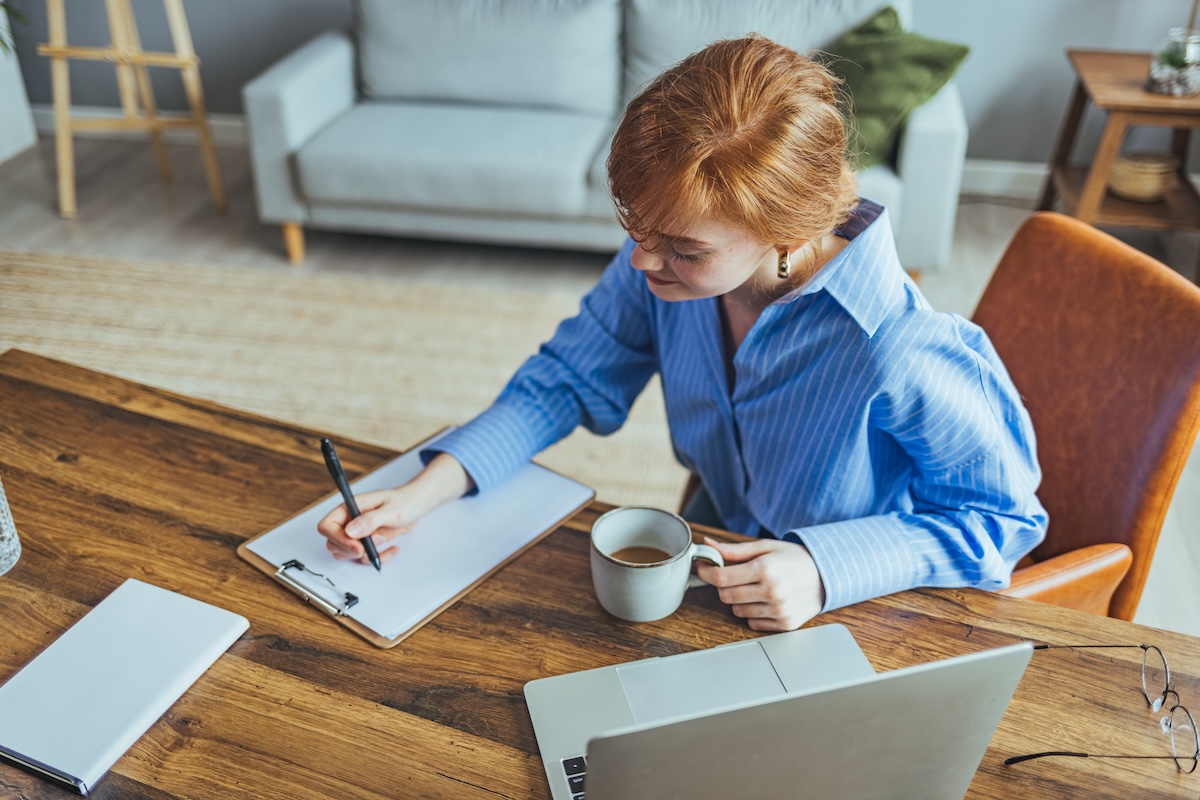 A woman with red hair writes a list on a pen an paper with a laptop nearby.