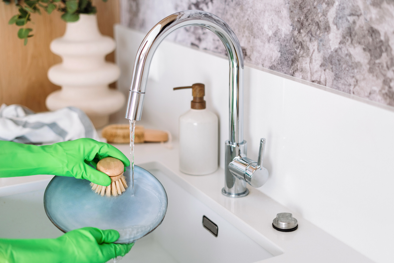 A person wearing green dish gloves and using a small brush to wash a plate in a ktichen sink with the best dish soap dispenser option in the background.