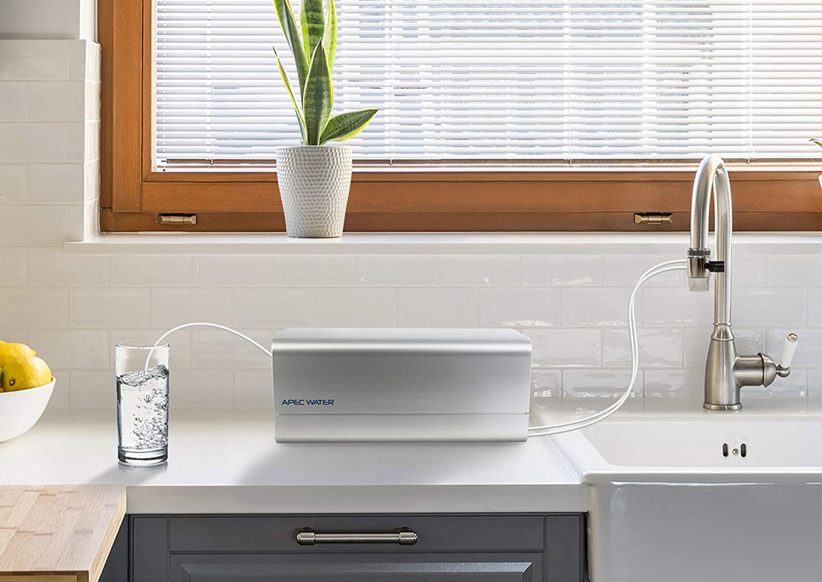 The Best Countertop Water Filter Option placed next to a kitchen sink and attached to its faucet on one side and being used to fill a glass of water on the other