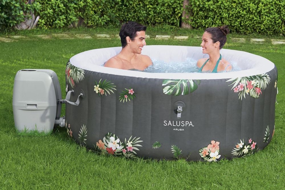 Two people sitting in Bestway SaluSpa Aruba AirJet Inflatable Hot Tub set up on green grass in a large yard.