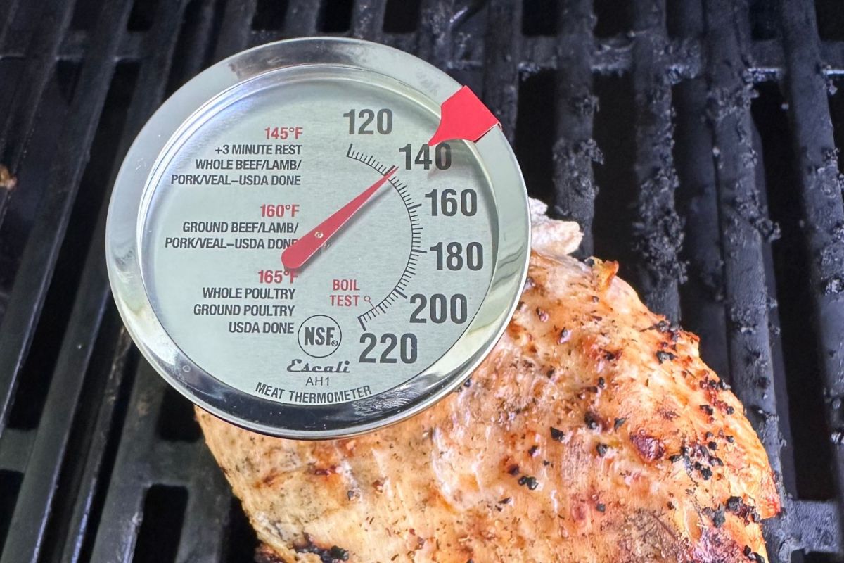 The Escali AH1 Oven-Safe Meat Thermometer inserted into a well-seasoned chicken breast cooking on a grill.
