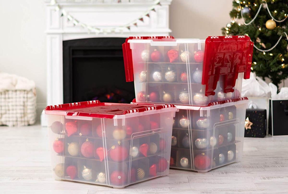Three of the Iris USA Holiday Ornament Wing-Lid Storage Boxes next to a tree and fireplace and full of red, silver, and gold ornaments.