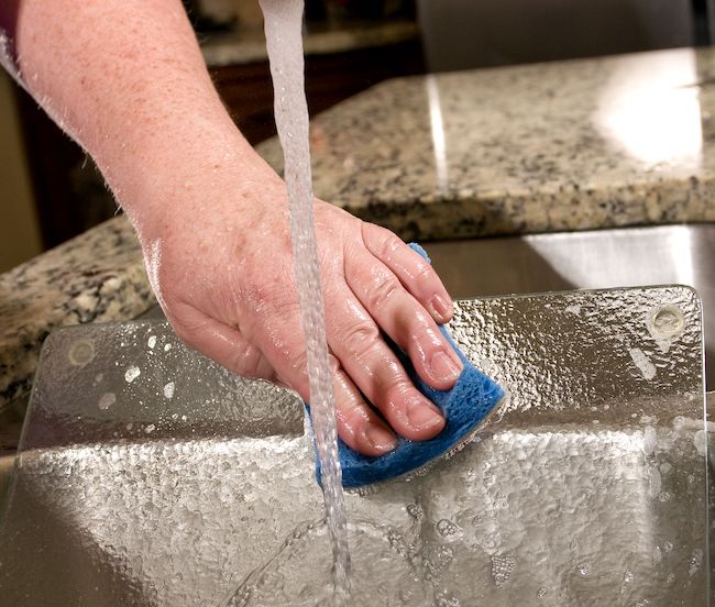 Cleaning a glass cutting board in her kitchen sink with a soapy sponge