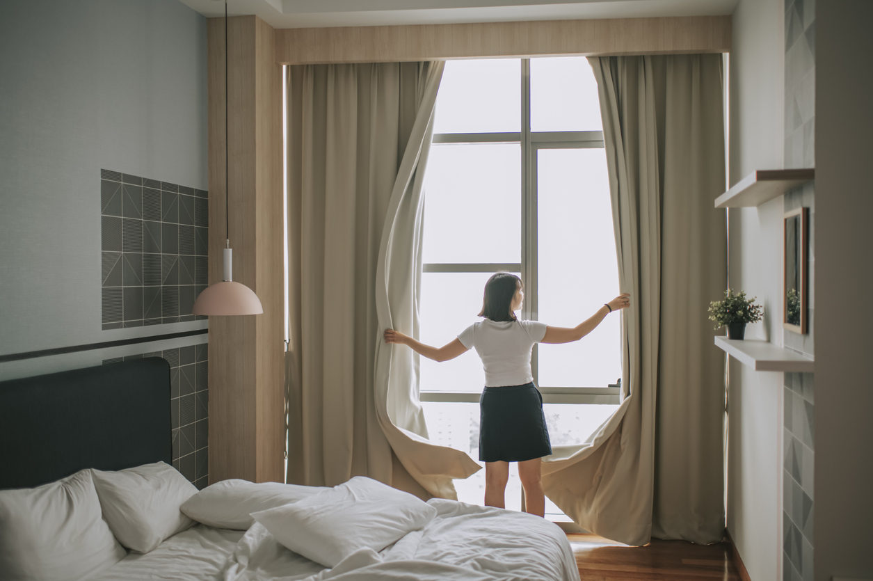 Woman opening thick curtains in bedroom, letting in morning light.