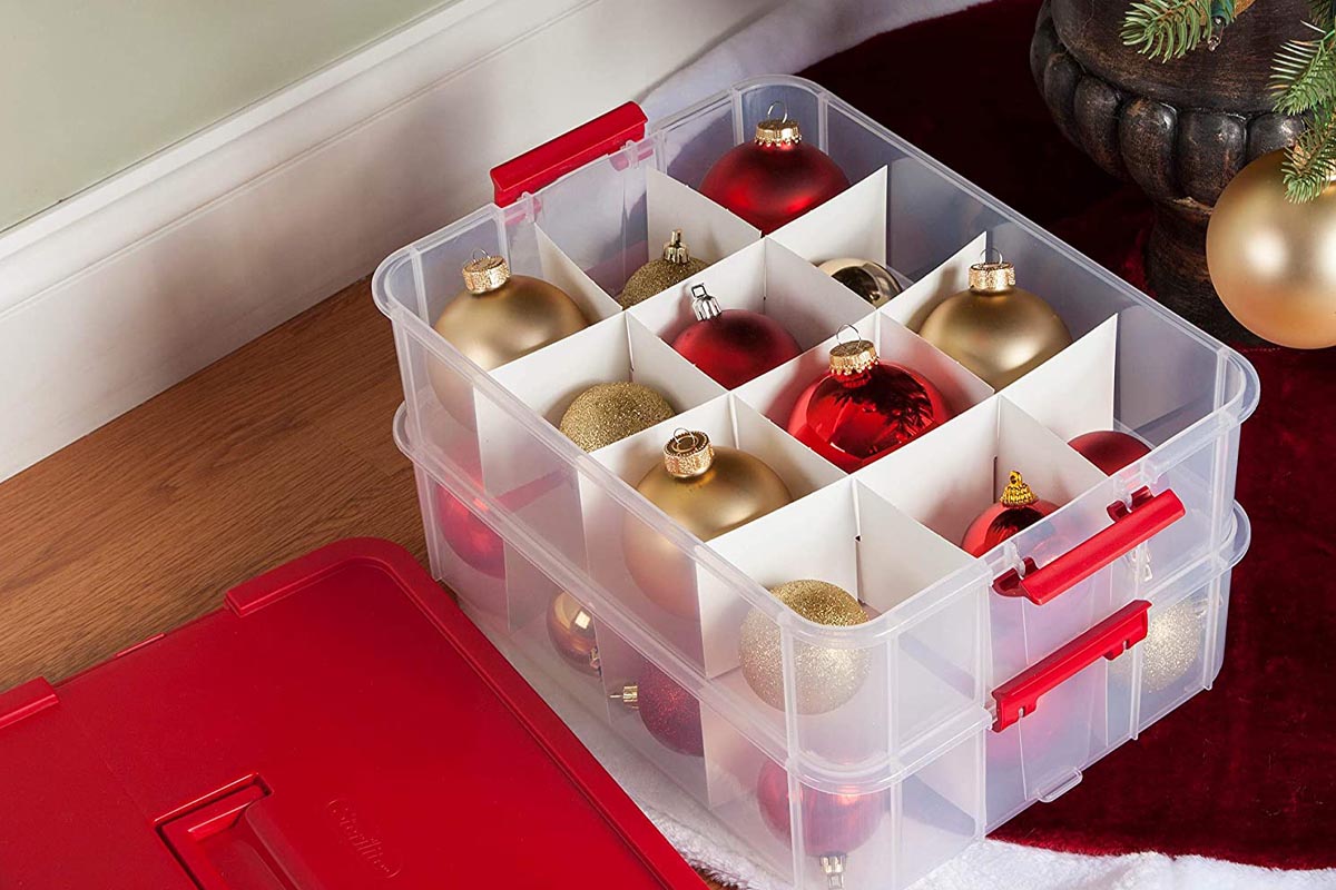 The Sterilite Christmas Ornament Storage Case on the floor next to a Christmas tree with its lid off to show red and gold ornaments.