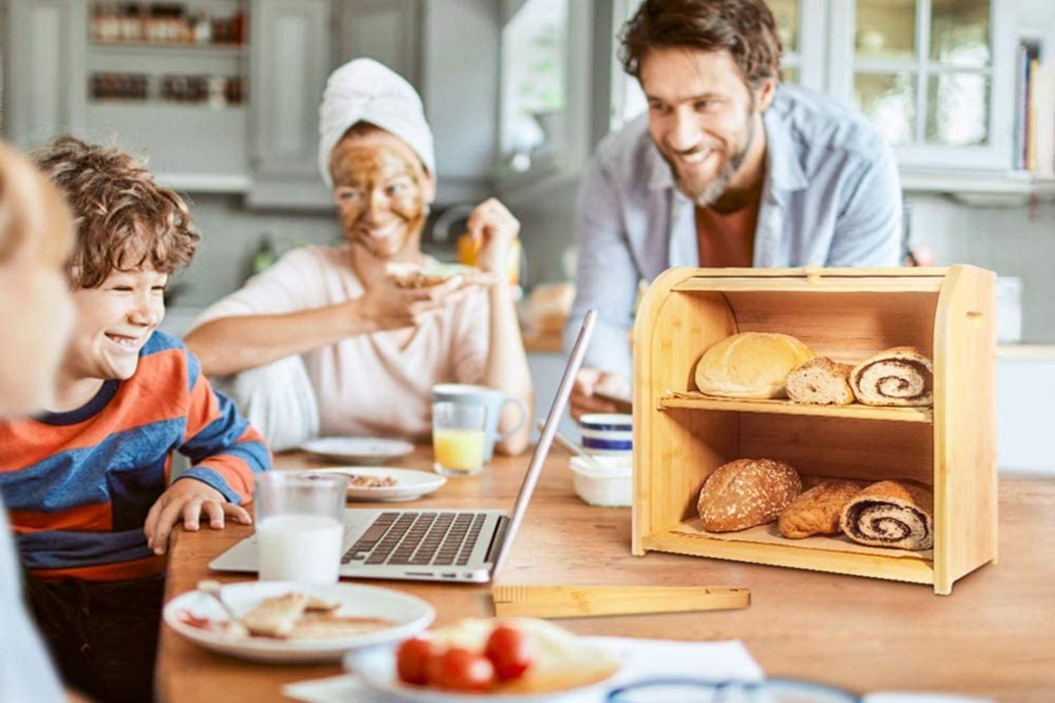 A family gathered at a kitchen counter with a laptop, breakfast items, and a bread box full of bread.