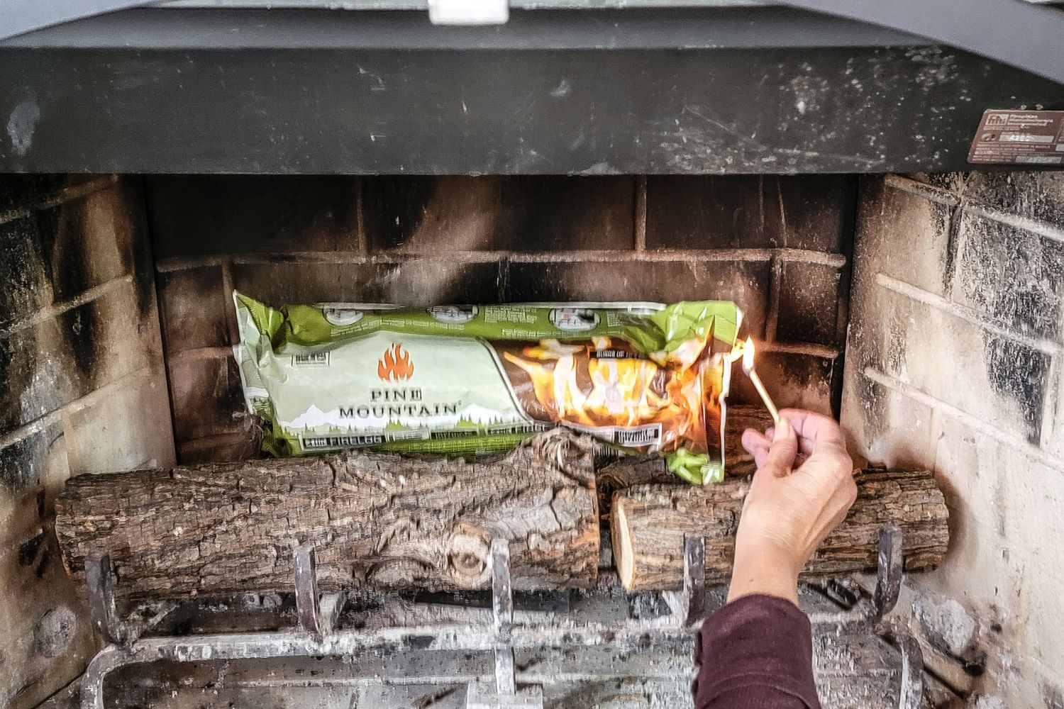 A person bringing a lit match to the end of a Pine Mountain fire log in a fireplace.
