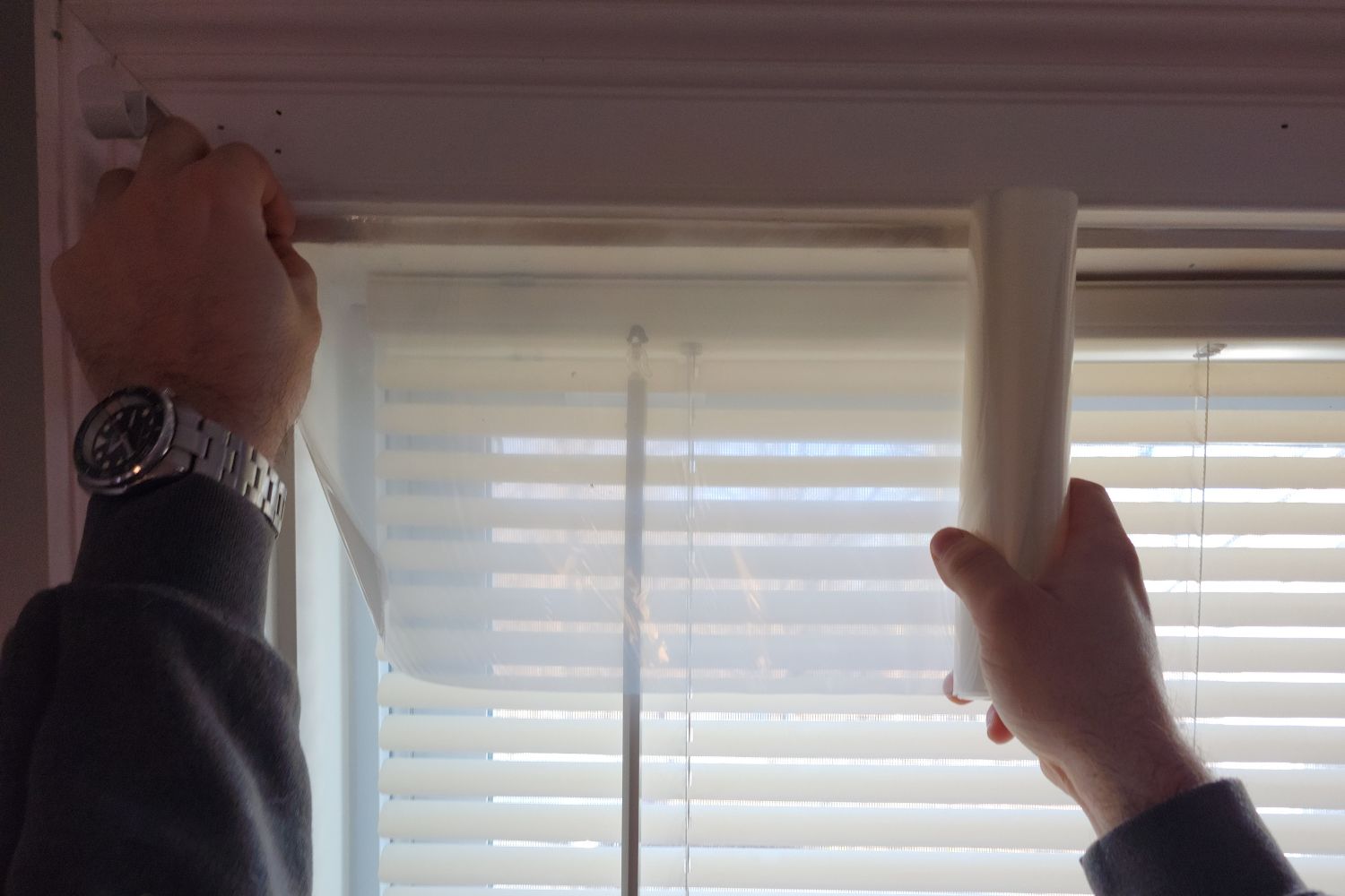 A person unrolling the best window insulation kit over a window with white mini blinds.