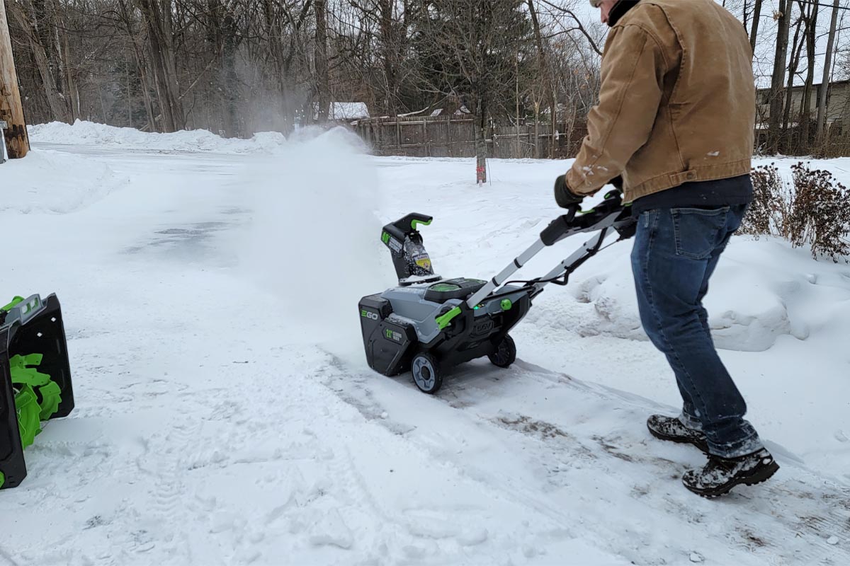 A person using an Ego cordless snow blower to clear snow from a fully covered driveway.