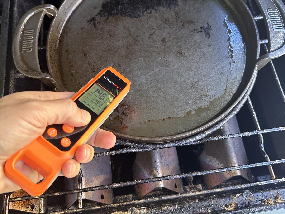 A person testing the surface temperature of a cast iron pan on a grill using the ThermoPro TP420 Meat Thermometer.