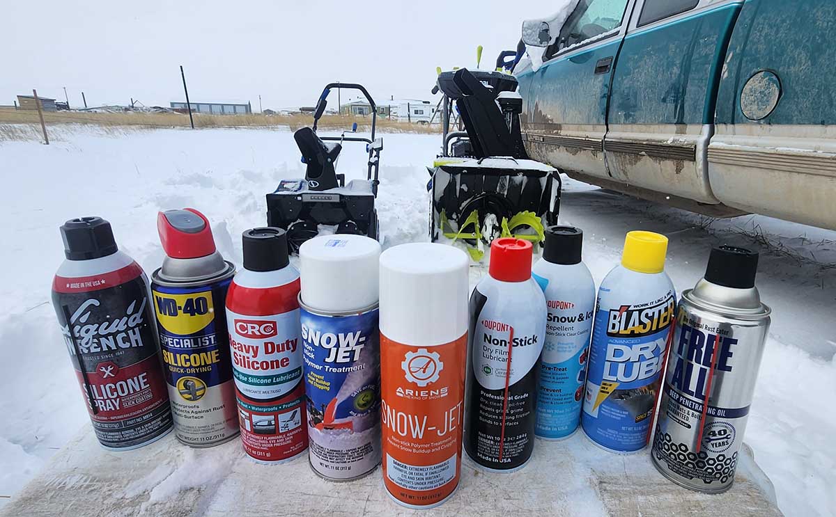 A group of the best tested nonstick sprays for snow blowers on a snowy surface with two snow blowers in the background.