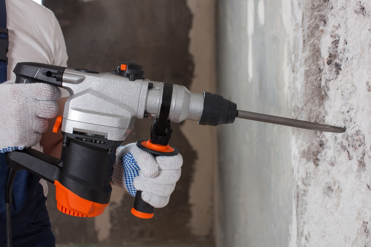 A home renovator using a silver and orange hammer drill in a cement wall.