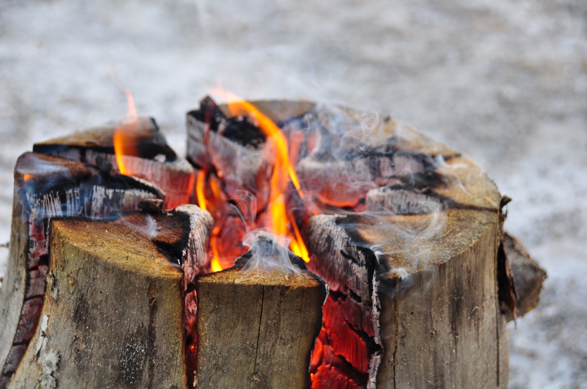 Outdoor tree stump cut in a crosshatch pattern with a fire blazing inside; light snow surrounds it.