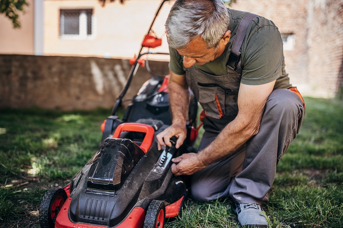 A man in work overalls repairs a strained electric lawn mower.