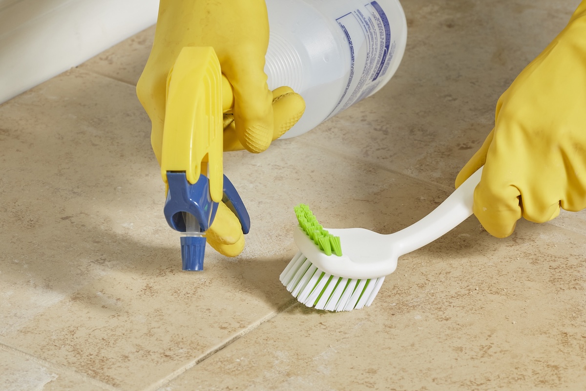 Person wearing yellow gloves sprays tile grout and scrubs with scrub brush.