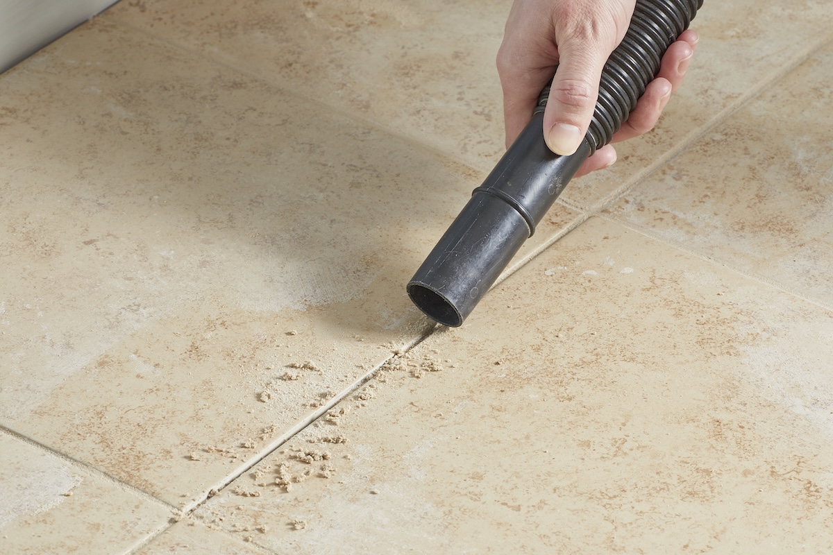 Person uses a vacuum attachment to suck grout dust off tile floor.