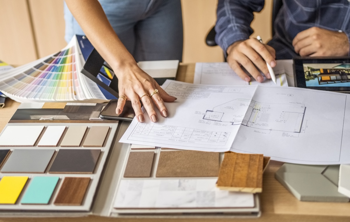 Cropped photo of two people's hands with blueprints, kitchen tile and color swatch samples on a table.