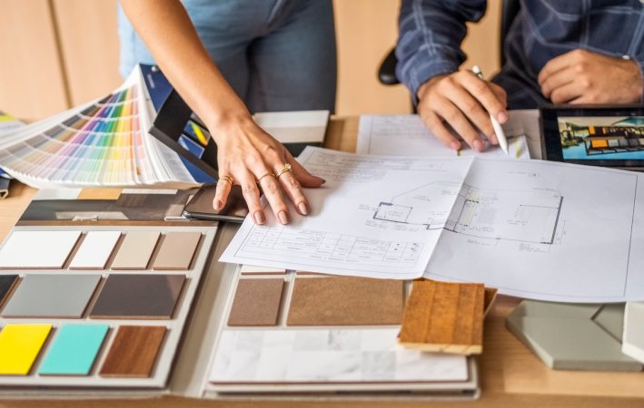 Cropped photo of two people's hands with blueprints, kitchen tile and color swatch samples on a table.
