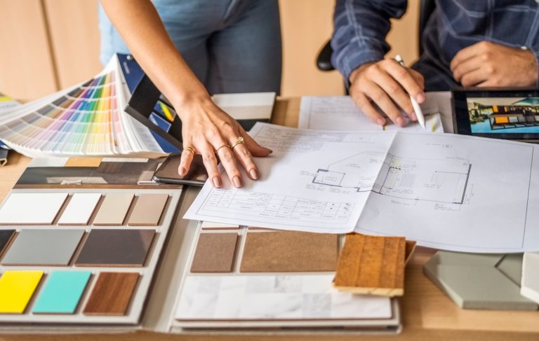 Cropped photo of two people's hands with blueprints, kitchen tile and color swatch samples on a table.