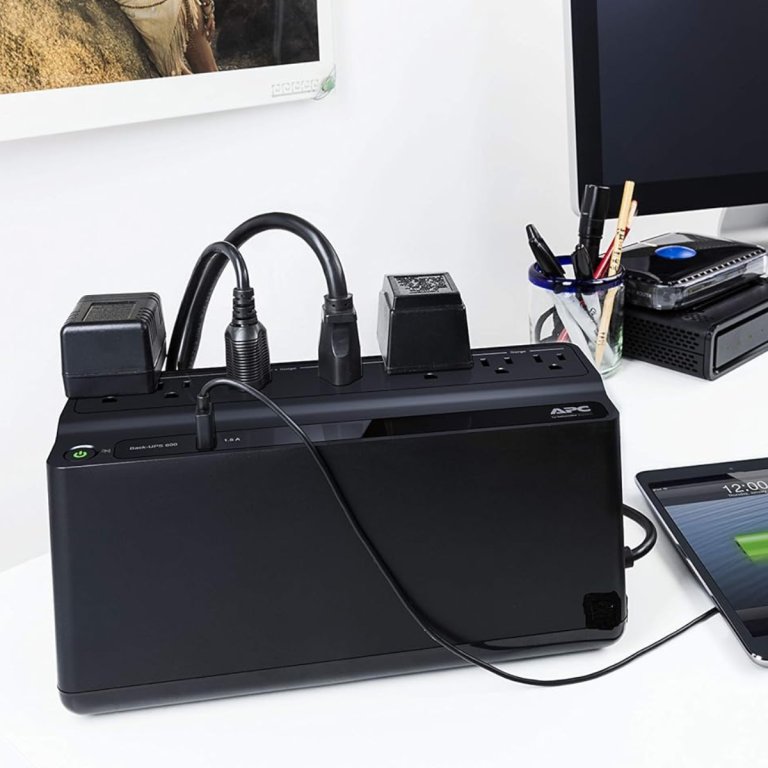 APC UPS Battery Backup & Surge Protector on a desktop with an iPad, computer, and other electronics plugged in.