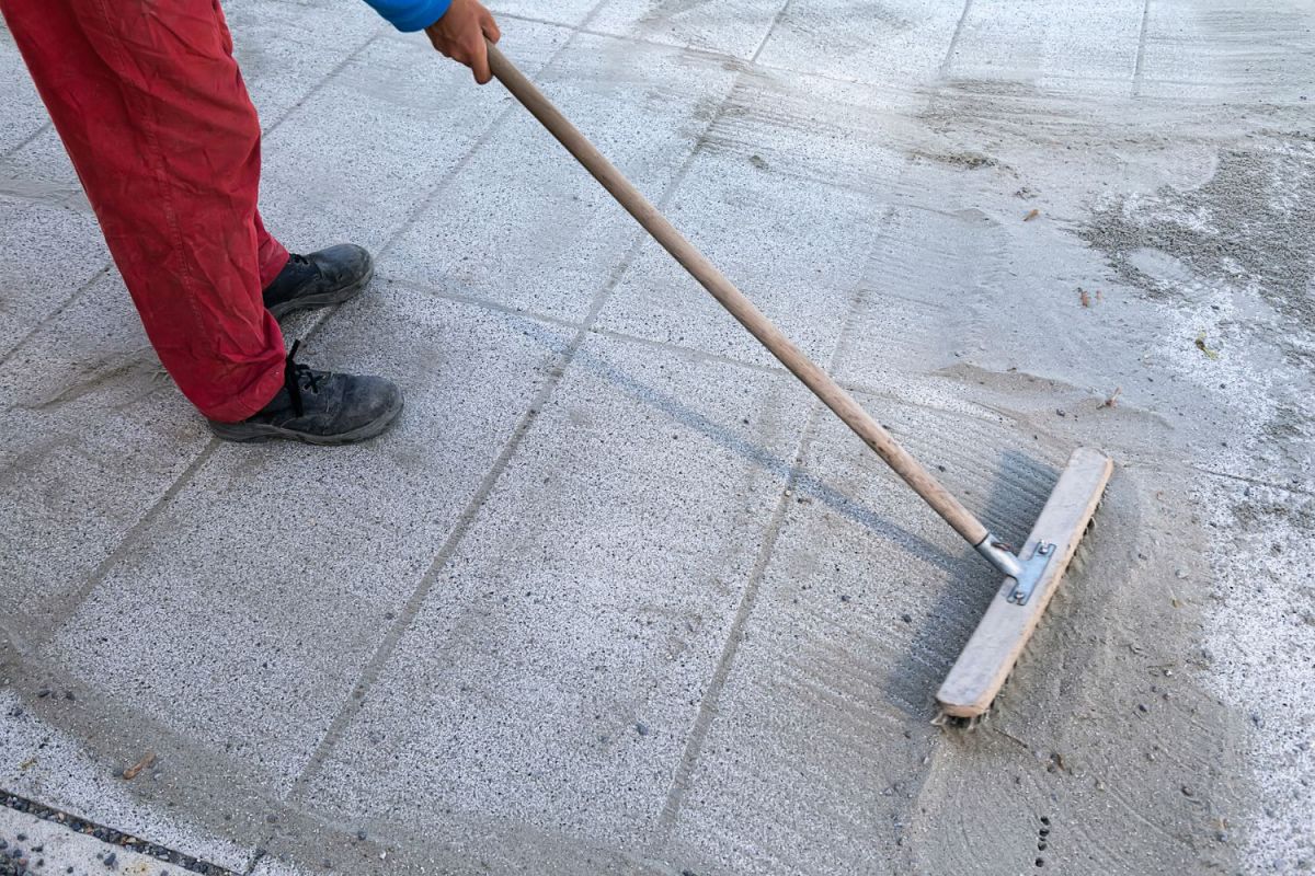 A person using a push broom to distribute the best polymeric sand between cement patio pavers.