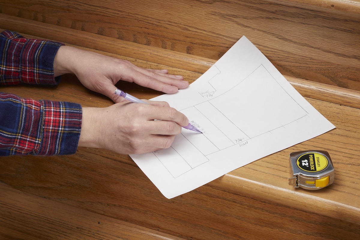 Woman sketching stair layout on one of the stair's steps, tape measure nearby.