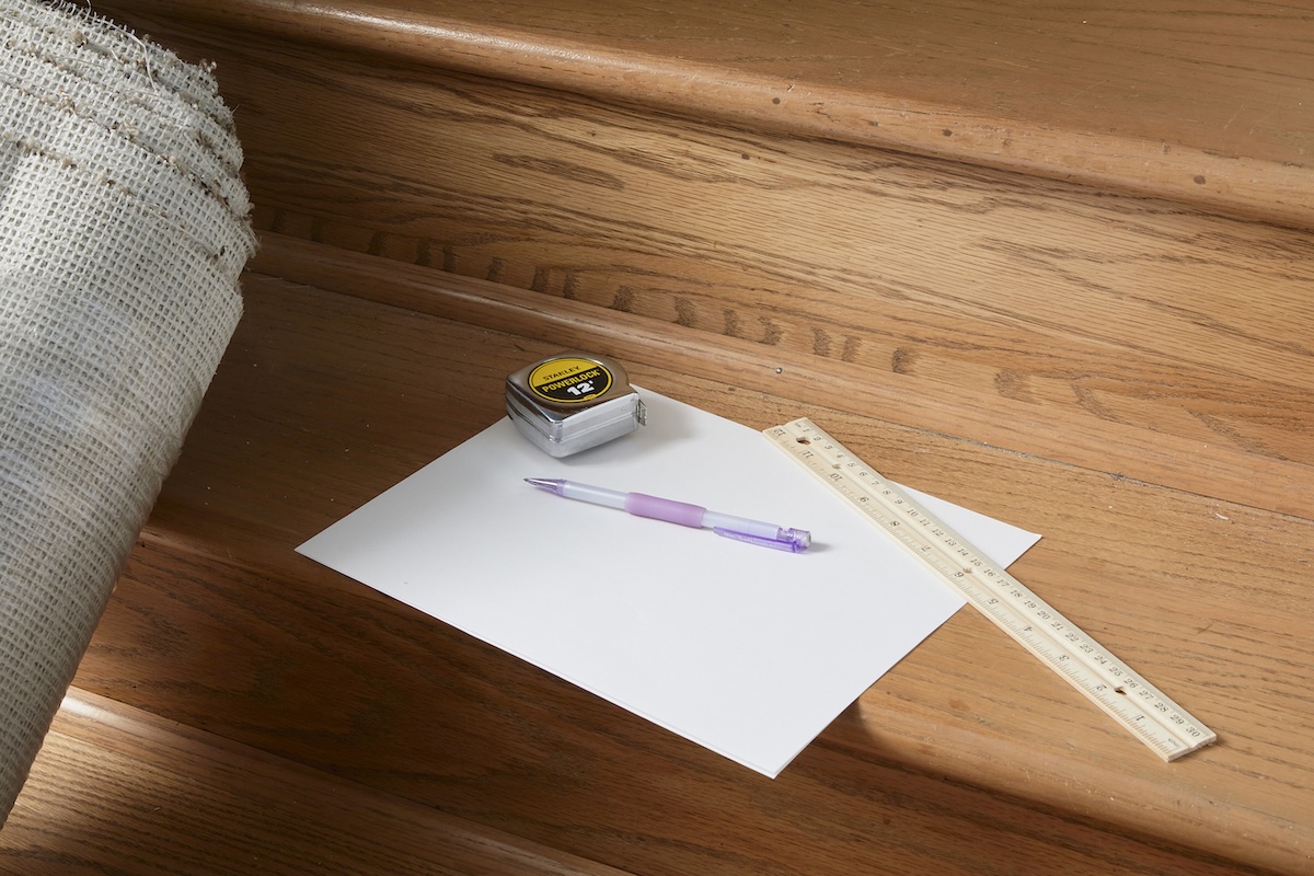 A pencil, piece of paper, tape measure, and ruler lay next to a rolled-up piece of carpet on a staircase.