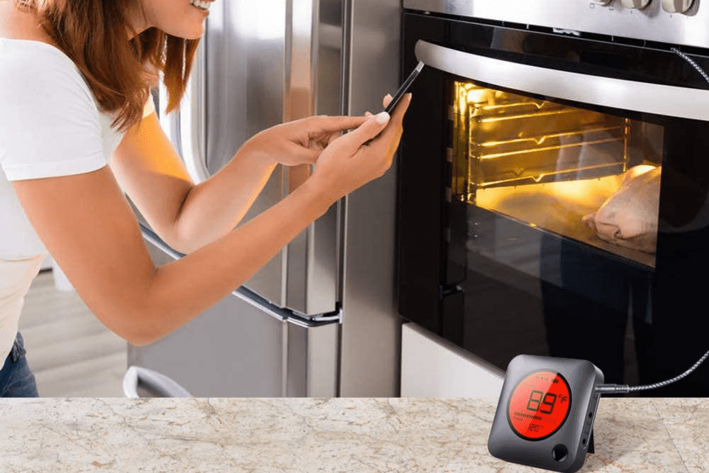 A person kneeling in front of an oven while looking at their phone with the best wireless meat thermometer in the foreground.