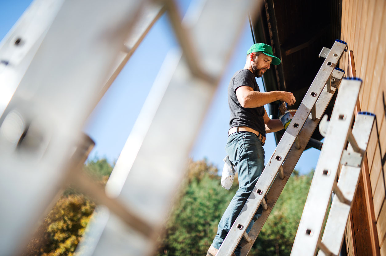 A worker stands on a ladder to paint the exterior of a house. 