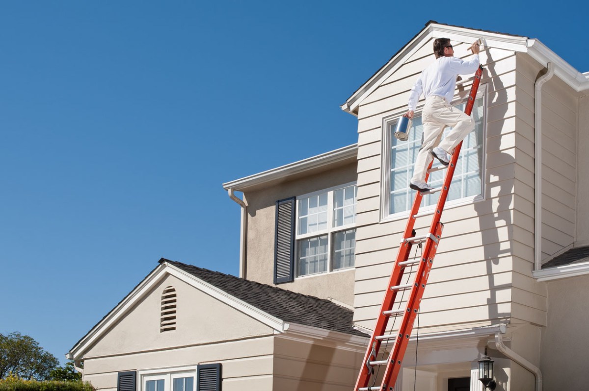 A worker stands on a ladder to paint the exterior of a house.