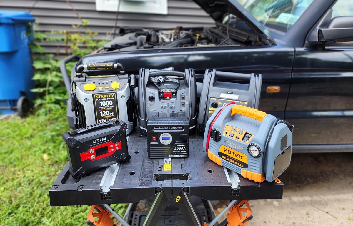 A group of the best jump starters with air compressors options on a work table next to a car with an open hood